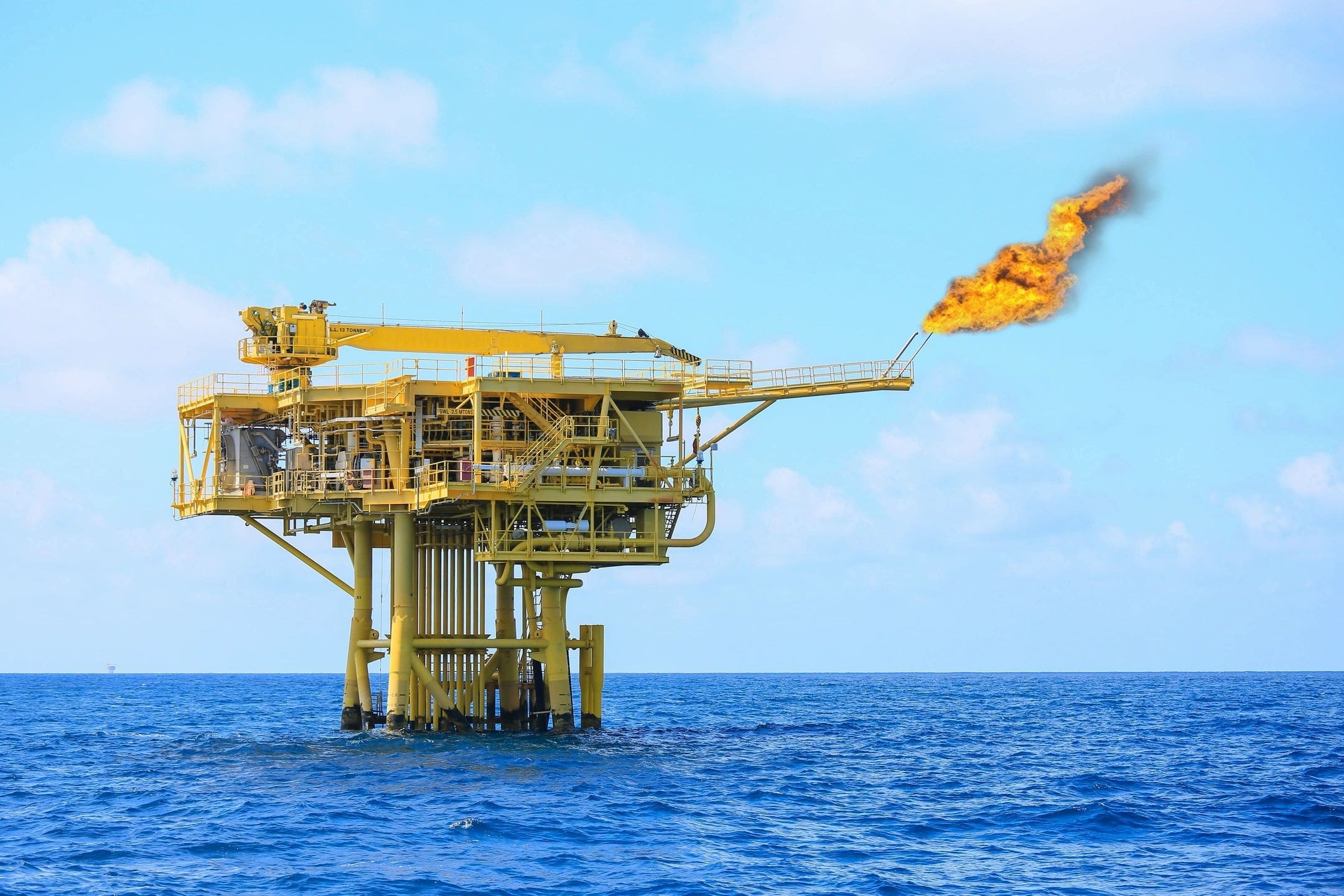 A yellow and white crane flying over an oil rig.